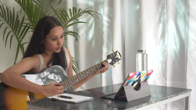 A focused woman is playing acoustic guitar and watching an online course on laptop tablet.Beautiful Indian girl learning guitar online using notepad laptop.Concept of online classes,practicing at home