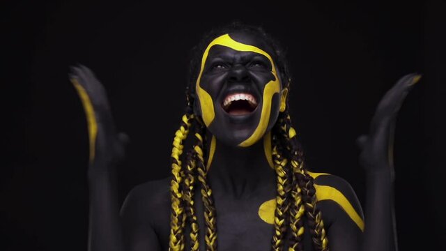 Face art. Screaming snarling wild woman with black and yellow body paint. Young african girl with colorful bodypaint. An amazing afro american model with yellow makeup.