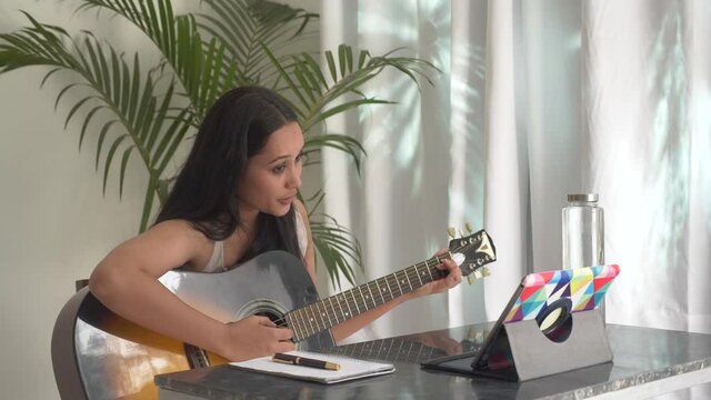 Beautiful Indian girl watching online guitar lesson and practicing music.Young Asian woman playing guitar at home.Aspiring musician learning to play musical instrument using laptop tablet.