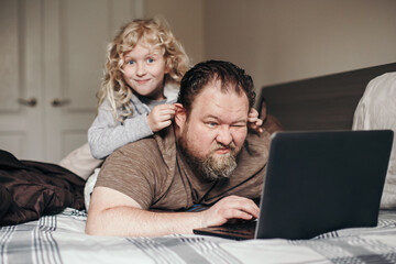 Work from home with kids children. Father working on laptop in bedroom with child daughter on his back. Funny candid family moments. New normal during coronavirus quarantine pandemic shutdown. - 414523942