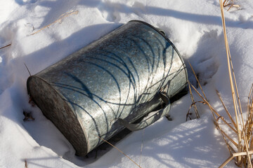 Gardening galvanized metal watering can on the snow in a winter garden. Geometric grass shadows. Selective focus.