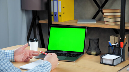 Businessman at home office sitting on chair at table with pen in hand tells with green screen laptop computer. Business man communicates employees company by online remote conference webcam video call