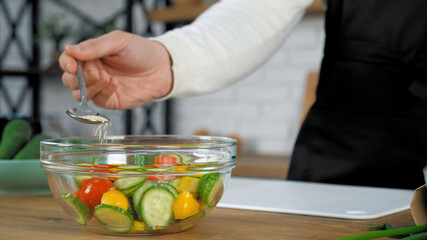 Hand professional chef in black apron standing near kitchen table sprinkle sesame seeds on cooked vegetarian salad in glass bowl. Man preparing delicious healthy vegan salad for breakfast or dinner