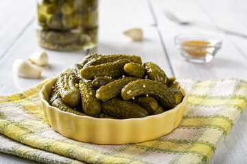 Delicious pickled cornichons in a yellow ceramic bowl over checkered napkin on a table. Whole green...
