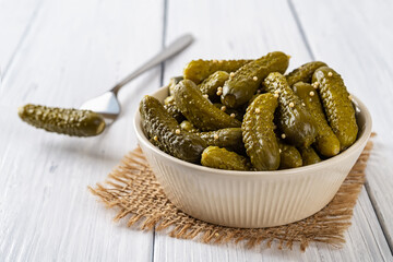 Crunchy pickled cornichons in a beige ceramic bowl and one on a fork on a white wood table. Whole...