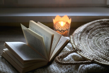 Open book, woven basket, crochet blanket and lit candle. Cozy details at home. Selective focus.