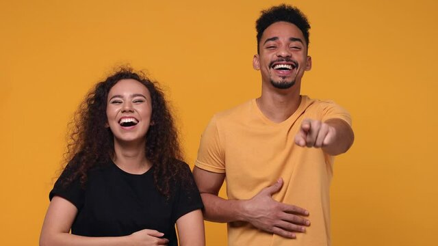 Excited laughing young couple two friends african american man woman 20s in basic t-shirt posing isolated on yellow background studio. People lifestyle concept. Pointing index fingers camera on you
