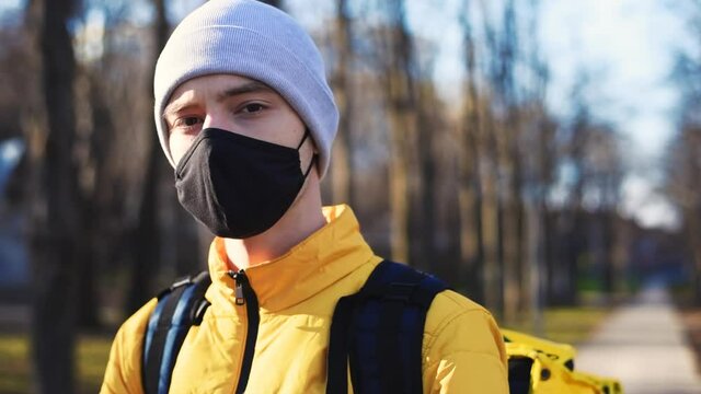 Food delivery man in a park looking into camera. Black medical mask, yellow backpack and jacket. Winter