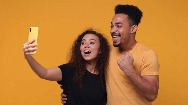 Funny couple friends african american man woman isolated on yellow background studio. People lifestyle concept. Doing selfie shot on mobile phone hugging showing thumb up biceps muscles okay gesture