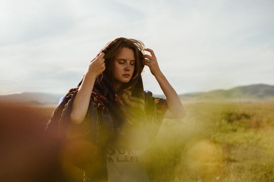 In the steppe grass, a girl straightens a cape on her head with sadness covering her eyes against a backdrop of hills in the distance. High quality photo