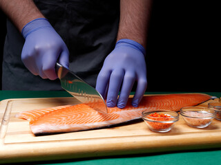 A young chef cuts a fresh raw salmon fillet and spices lie nearby on a wooden cutting board....