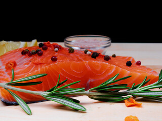 Close-up of slice of red fresh raw salmon fillet with pepper and spices and rosemary next to it. lie on a wooden cutting board. Focus on rosemary and salmon. Healthy food concept