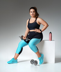 Fototapeta premium Sporty girl resting after workout with dumbbells. Photo of model with curvy figure in fashionable sportswear on grey background. Sports motivation and healthy lifestyle
