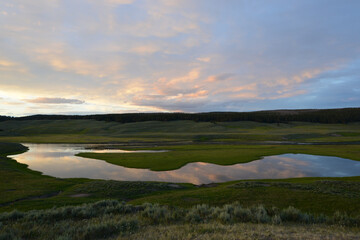 View of a river flowing through a meadow at Yellowstone National Park at sunset, with reflections of colorful clouds in the water