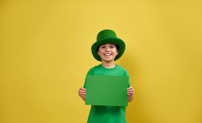 Smiling boy wearing green Irish leprechaun hat poses wit a sheet of green paper in his hands. Celebrating Saint Patrick's Day concept. Yellow background. Copy space.
