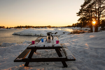 Outdoors coffee break in the winter landscape by the sea during sunset. Thermos and mugs on picnic table covered in snow. Photo taken in the archipelago outside Oskarshamn, Sweden