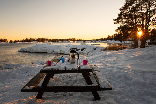 Outdoors coffee break in the winter landscape by the sea during sunset. Thermos and mugs on picnic table covered in snow. Photo taken in the archipelago outside Oskarshamn, Sweden