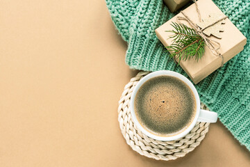A cup of coffee and Christmas gifts on green sweater on beige background, top view, copy space
