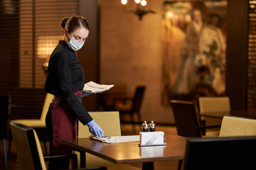 Competent waitress in protective mask preparing table