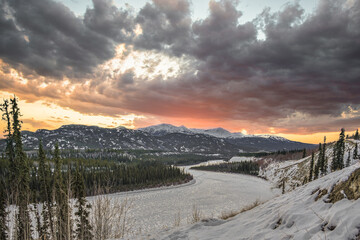 A stunning winter season view of the Yukon River in northern Canada with stormy, snowy looking clouds above at pink, orange sunset hour with snow capped mountains. 