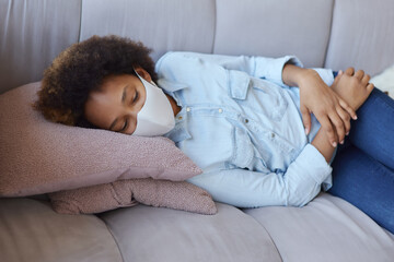 Mixed race teen girl in casual clothes wearing protective face mask relaxing, lying on the couch while spending time at home during quarantine