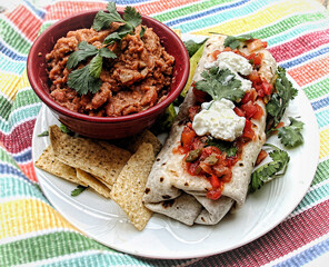 Plate of a burrito with salsa and sour cream and a bowl of refried beans, set on a colorful placemat.