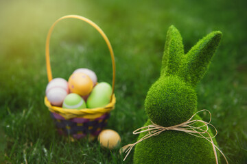 Close up Easter bunny rabbit statuette and basket with easter eggs on the green grass lawn background. Easter egg hunt in the garden. Selective focus, copy space.