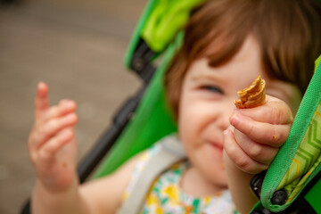 A cute, blue-eyed, brown-haired baby girl sitting in a green pushchair holding a piece of food and looking at the camera