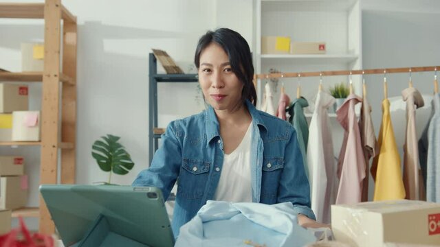 Young Asia lady fashion designer using mobile phone receiving purchase order and showing clothes recording video live streaming online at shop. Small business owner, online market delivery concept.