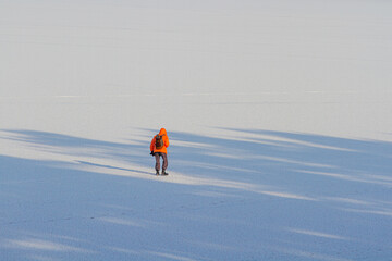 A man in an orange jacket walks in the snow on a frozen lake.