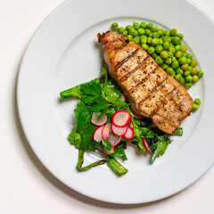 Grilled pork steak with green vegetables and peas on white plate view from directly above