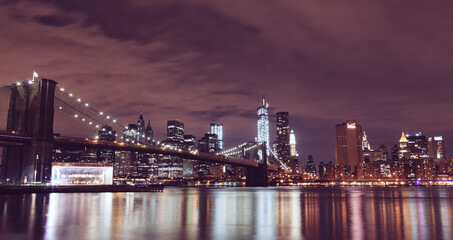 Illuminated Brooklyn Bridge And Buildings By River Against Sky At Night in New York City