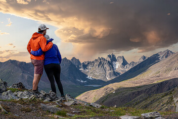 Amazing jagged mountain view in northern Canada, Yukon Tombstone Territorial Park with couple hugging. Orange and purple clothing with stormy looking sky in background. 