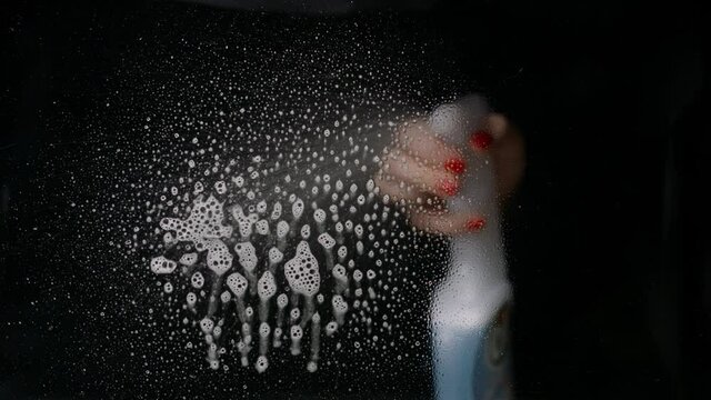 Female hand sprays cleaning agent onto the glass. Foaming bubbles comes down