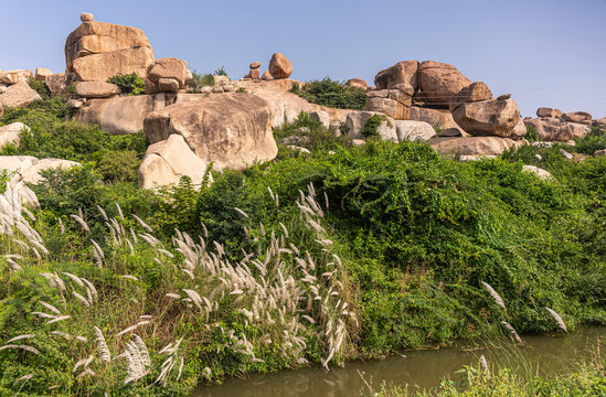 Hampi, Karnataka, India - November 9, 2013: Brown rock boulder landscape against blue sky above green foliage and creek near Tungabhadra Bukka Aqueduct.