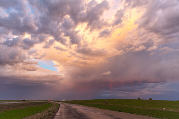 A flat landscape highway seen before a cloudy storm approaching with pink, orange dark clouds, shining sun and dark country, rural land below. Great for sky replacement for editing photos or drawing. 