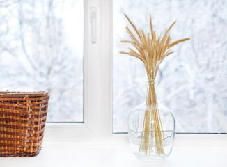 Wheat ears in a glass vase with wicker box on a winter windowsill. Eco interior concept.
