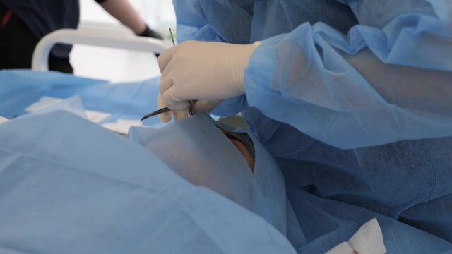 Hands of surgeon doctor before surgical operation preparing patient for facial cosmetology surgery. Surgeon in blue medical uniform and gloves doing plastic operation on eyelids with scalpel.