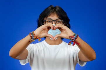 A young man with hands in the shape of a heart wears a medical mask and a white shirt on a blue background.
