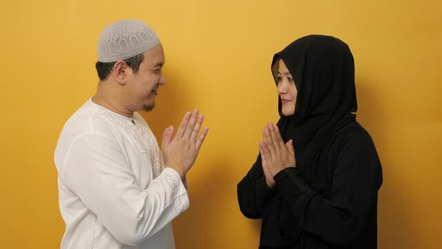 Happy Asian muslim couple doing greeting gesture, apologize and forgiving each other on eid mubarak tradition, against yellow