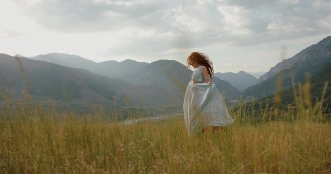 Girl in light dress with red hair is walking on top of a spring mountain with scenic view. Woman in search for inspiration - freedom concept 4k footage