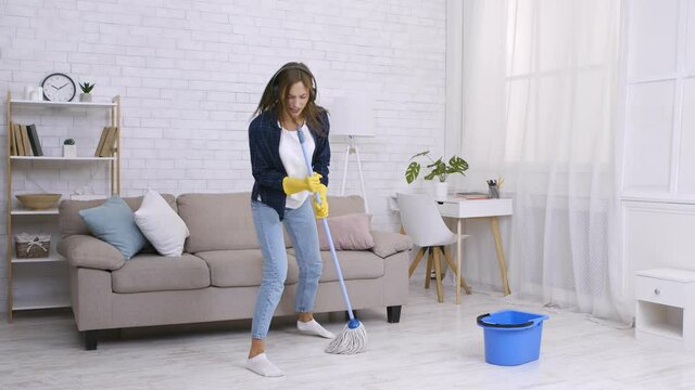 Spring cleaning is fun. Happy young woman dancing and singing into mop like mic, listening to music in headphones