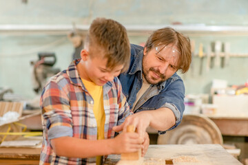 Father teaches to young boy to plan wood in a carpentry workshop