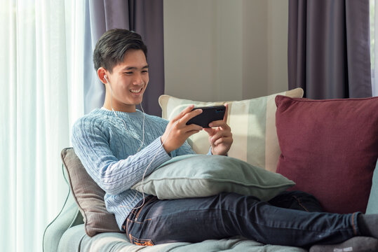 Happy Young Asian Man Wearing Headphones Playing Games At Home On The Sofa.