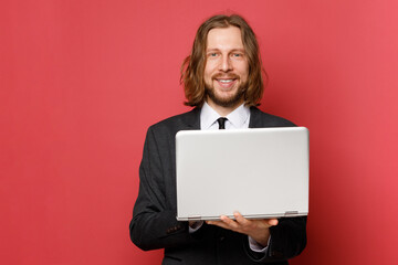 Attractive smiling adult businessman with a beard and long hair in a formal suit with a laptop on a red background. 