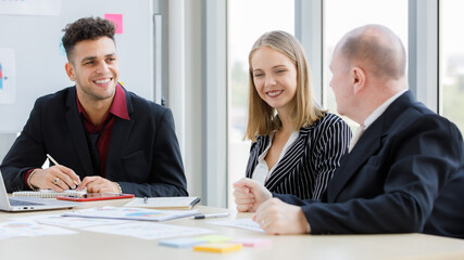 2 Adults caucasian businessmen are talking. A focused businessman is holding pen and look at a teammate and have brown cup, book, laptop put on desk and whiteboard are back with more graph on paper