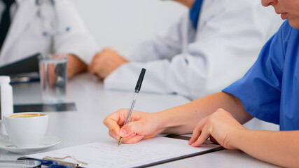Close up of nurse writing on clipboard while coworkers discussing in background analysing patients...