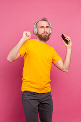 Handsome man in casual dancing with mobile phone and headphones isolated on pink background