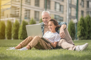 Son and dad sitting together on the grass with a laptop and looking contented