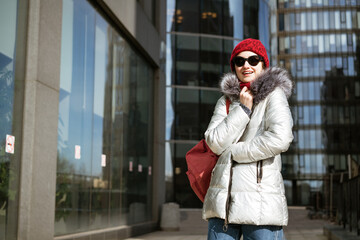 Happy young woman in sunglasses in a winter jacket with a backpack near the building. Travel concept